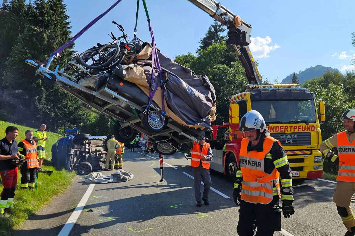 Ein Abschleppfahrzeug hebt einen umgekippten Anhänger mit Fahrrädern. Feuerwehrleute sichern die Unfallstelle auf einer Landstraße.