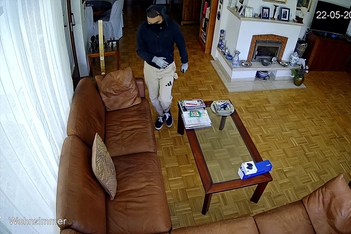 Eine maskierte Person betritt ein Wohnzimmer mit Ledercouch und Glastisch. Im Hintergrund ist ein Kamin zu sehen.