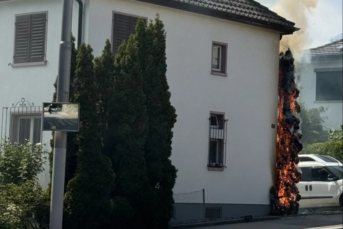 Ein Feuer breitet sich an der Außenwand eines weißen Gebäudes aus. Daneben steht ein geparktes Fahrzeug.