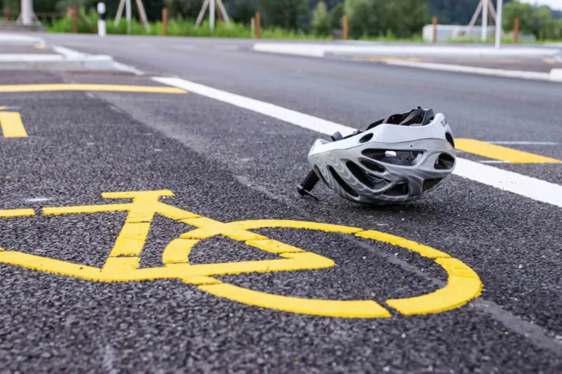 Ein grauer Fahrradhelm liegt auf einer Straße, die mit einer gelben Fahrradweg-Markierung versehen ist. Im Hintergrund sind weitere Fahrbahnmarkierungen und Begrenzungen zu sehen.