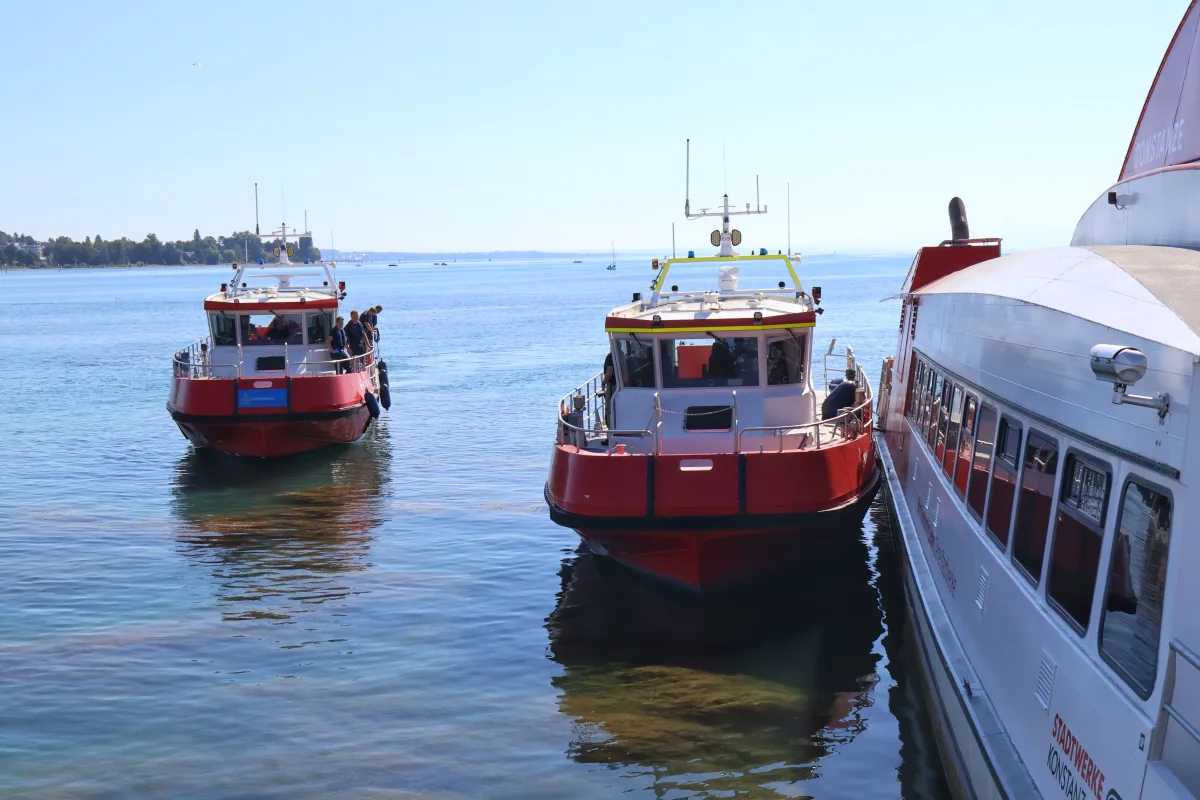 Zwei rote Boote der Polizei und ein größeres Schiff befinden sich auf einem ruhigen See unter klarem Himmel. Menschen sind an Bord der Boote erkennbar.