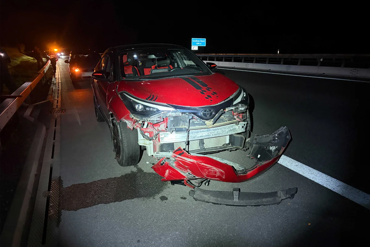 Ein roter PKW mit stark beschädigter Front steht auf einer abgegrenzten Autobahnspur bei Nacht. Das Auto zeigt einige Verformungen und abgelöste Teile.