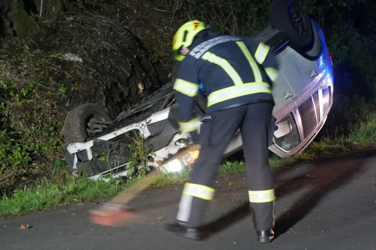 Ein Auto liegt bei Nacht auf dem Dach neben einer Landstraße. Ein Feuerwehrmann mit leuchtender Schutzkleidung ist vor Ort und arbeitet an der Unfallstelle.