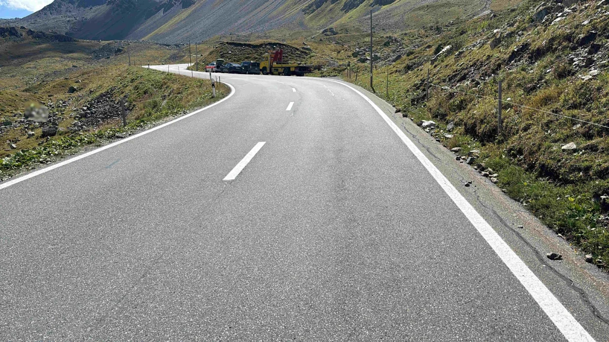 Eine kurvige Bergstraße wird von mehreren Fahrzeugen in der Ferne blockiert. Die Szene befindet sich in einer alpinen Umgebung mit steilen Hängen.