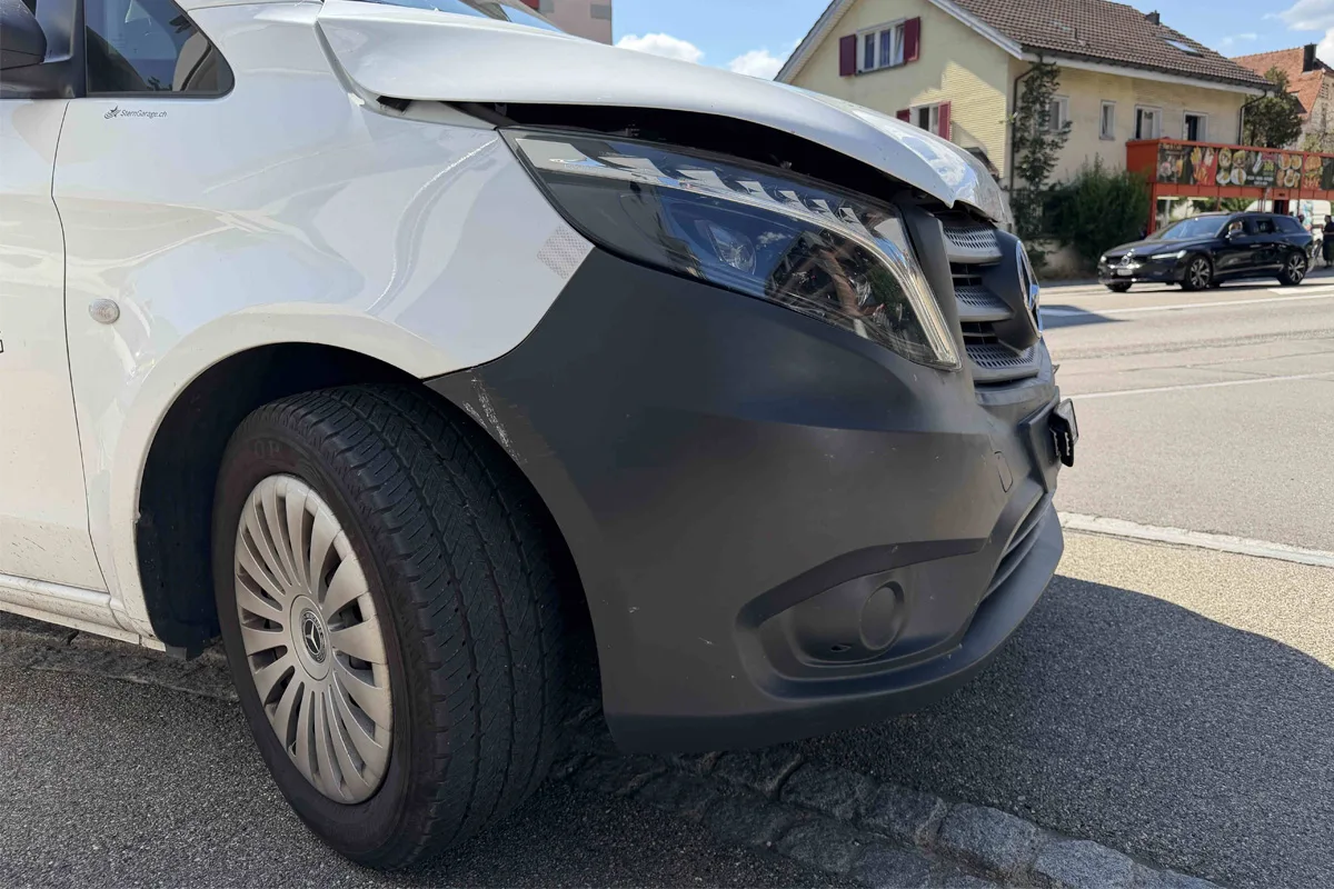 Ein weißer Mercedes-Van mit sichtbarem Frontschaden steht an einer Straßenecke. Im Hintergrund sind weitere Fahrzeuge und Wohnhäuser zu sehen.