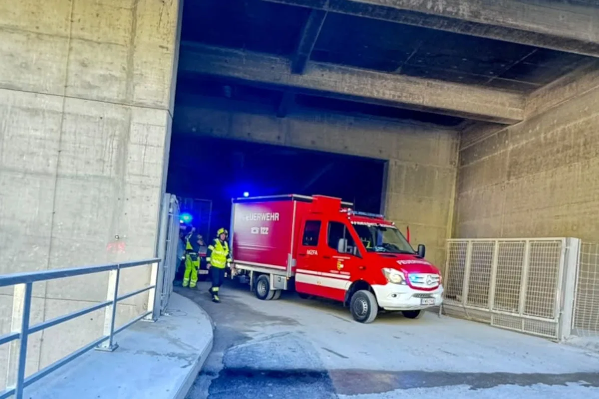 Ein rotes Feuerwehrfahrzeug verlässt einen Tunnel, begleitet von mehreren Feuerwehrleuten in gelben Westen. Der Tunnel und die Umgebung sind aus Beton.