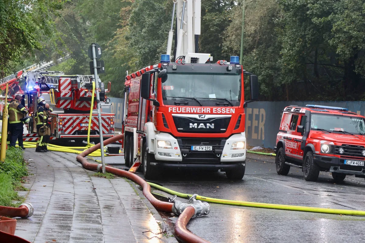 Ein Feuerwehrauto der Feuerwehr Essen steht mit ausgelegten Schläuchen auf einer nassen Straße. Im Hintergrund sind Einsatzkräfte aktiv.