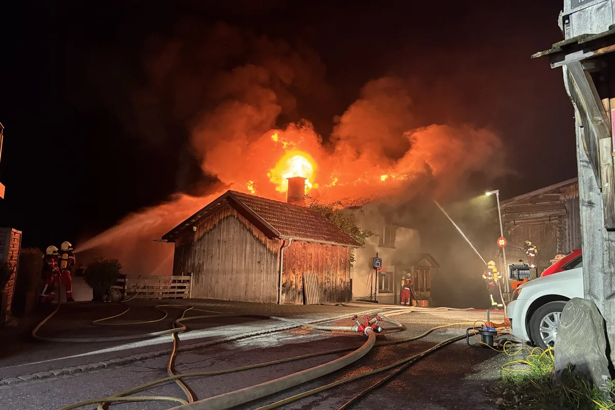 Ein Haus steht nachts im Vollbrand mit Flammen, die aus dem Dach schlagen, während Feuerwehrleute mit Schläuchen den Brand bekämpfen.