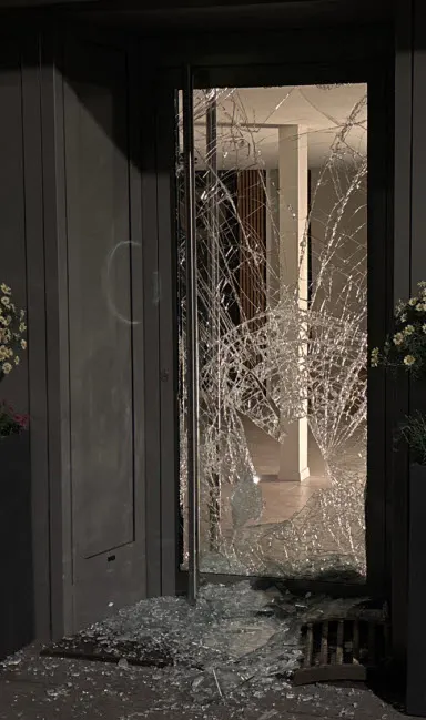 Das Bild zeigt eine Glastür, die stark beschädigt ist, mit Glasscherben am Boden in einem dunklen Eingangsbereich.