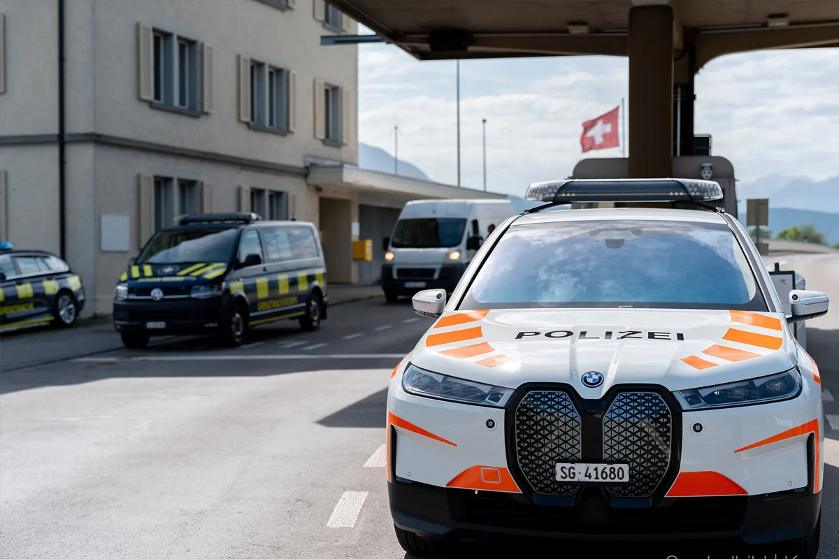 Im Vordergrund steht ein Schweizer Polizeiauto mit der Aufschrift 'Polizei' an einem Grenzposten. Dahinter sind weitere Fahrzeuge zu sehen, darunter ein Polizeivan und eine Schweizer Flagge im Hintergrund.