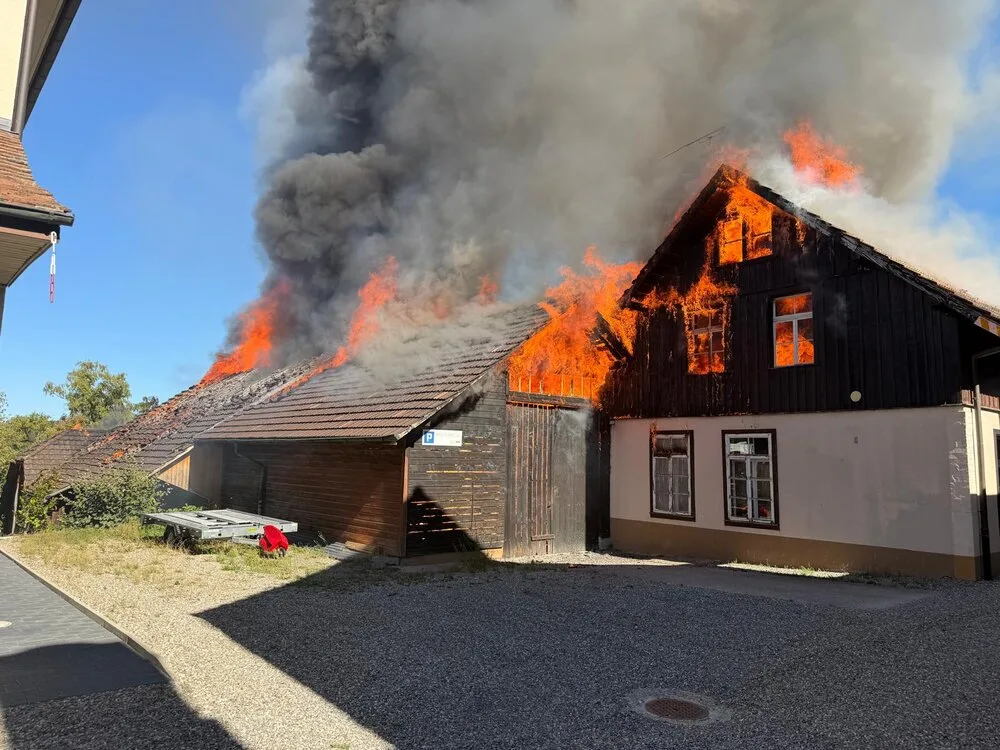 Ein Hausdach steht in Flammen, dichter Rauch steigt auf; das Gebäude ist stark vom Feuer betroffen.