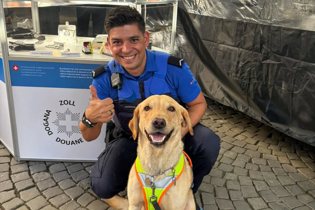 Ein Zollbeamter in blauer Uniform sitzt neben einem Hund mit Warnweste. Hinter ihnen sind beschlagnahmte Gegenstände ausgestellt.