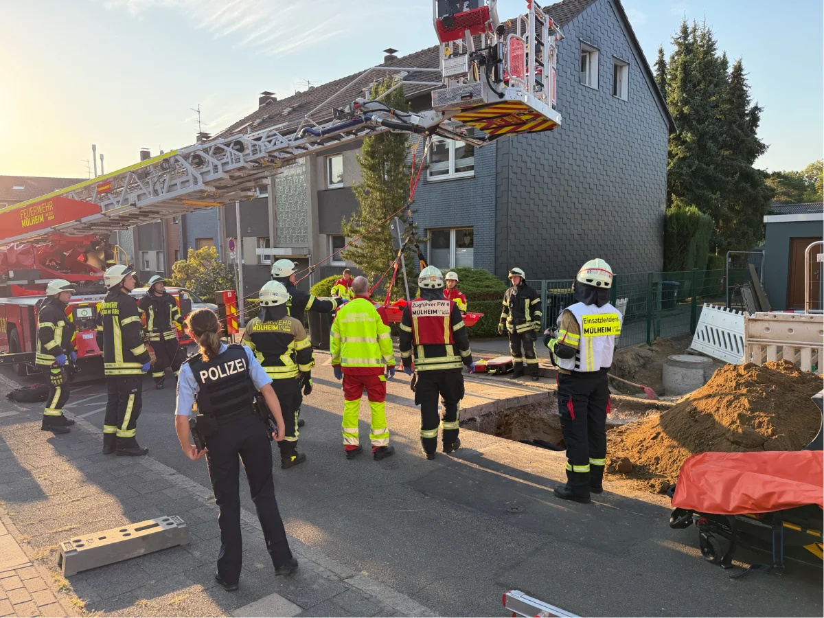 Feuerwehrleute und eine Polizistin stehen um einen Einsatzort, nahe eines Feuerwehrfahrzeugs und einer Baustelle. Ein Feuerwehrkran ist in der Luft, und es ist Abenddämmerung.