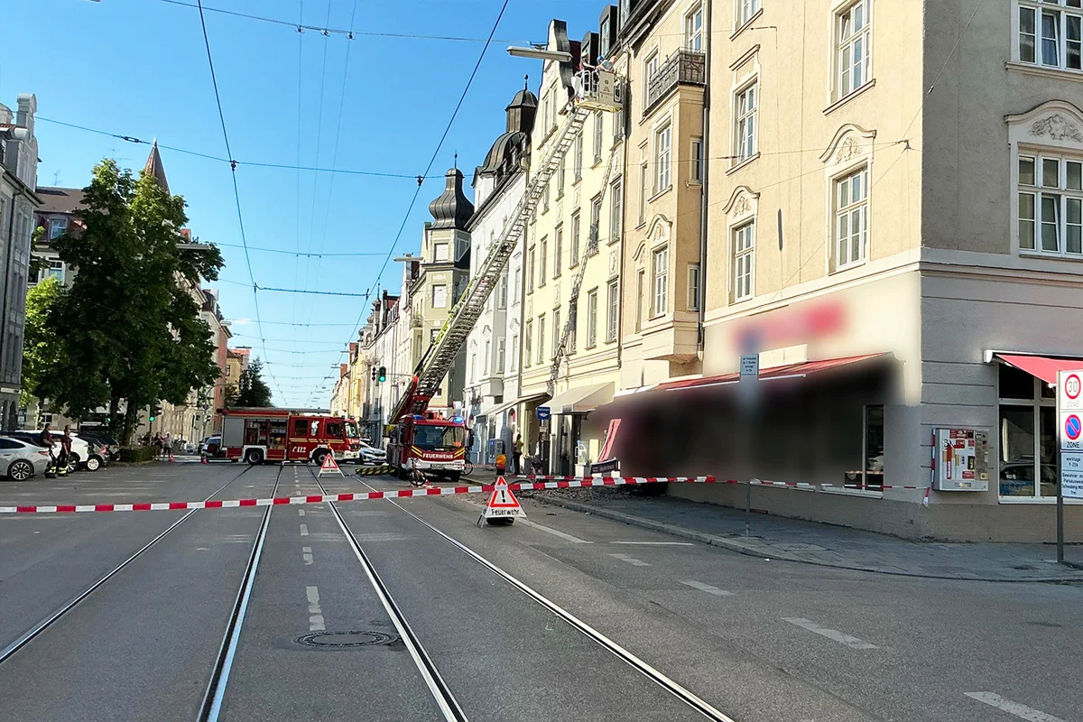 Mehrere Feuerwehrfahrzeuge stehen auf einer städtischen Straße, eines mit ausgefahrener Leiter in Richtung eines mehrstöckigen Wohnhauses. Absperrungen sind zu sehen.