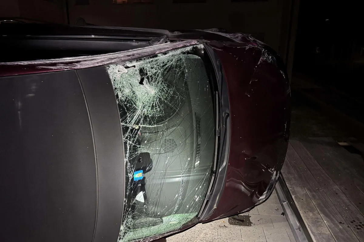 Ein Auto liegt seitlich auf dem Boden mit einer stark beschädigten Windschutzscheibe. Das Fahrzeug zeigt Spuren von erheblichem Aufprall.