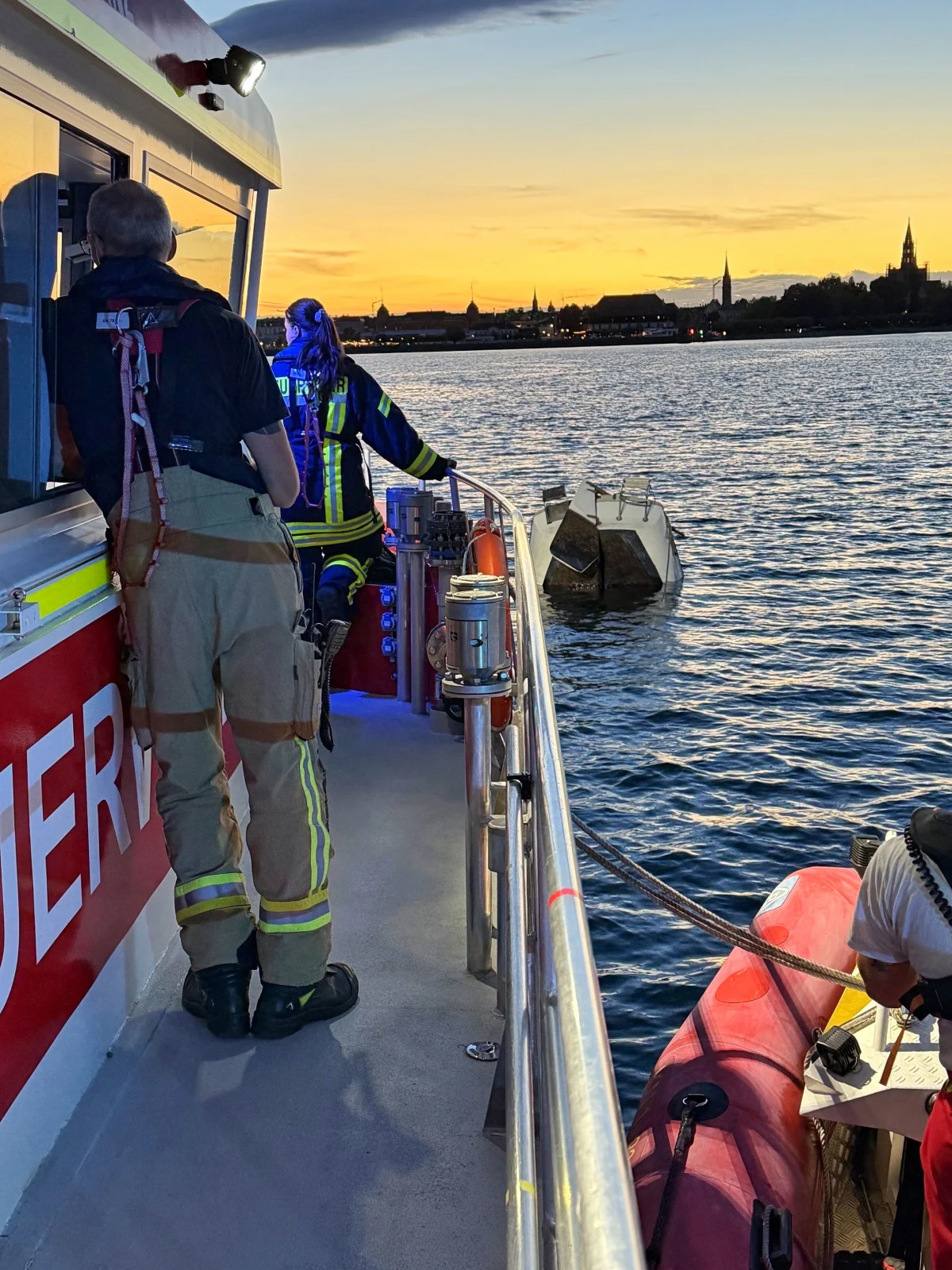Auf einem Boot sind Rettungskräfte unter einem farbenfrohen Sonnenuntergang auf einem See zu sehen. Ein kleines, teilweise untergetauchtes Boot befindet sich im Wasser.