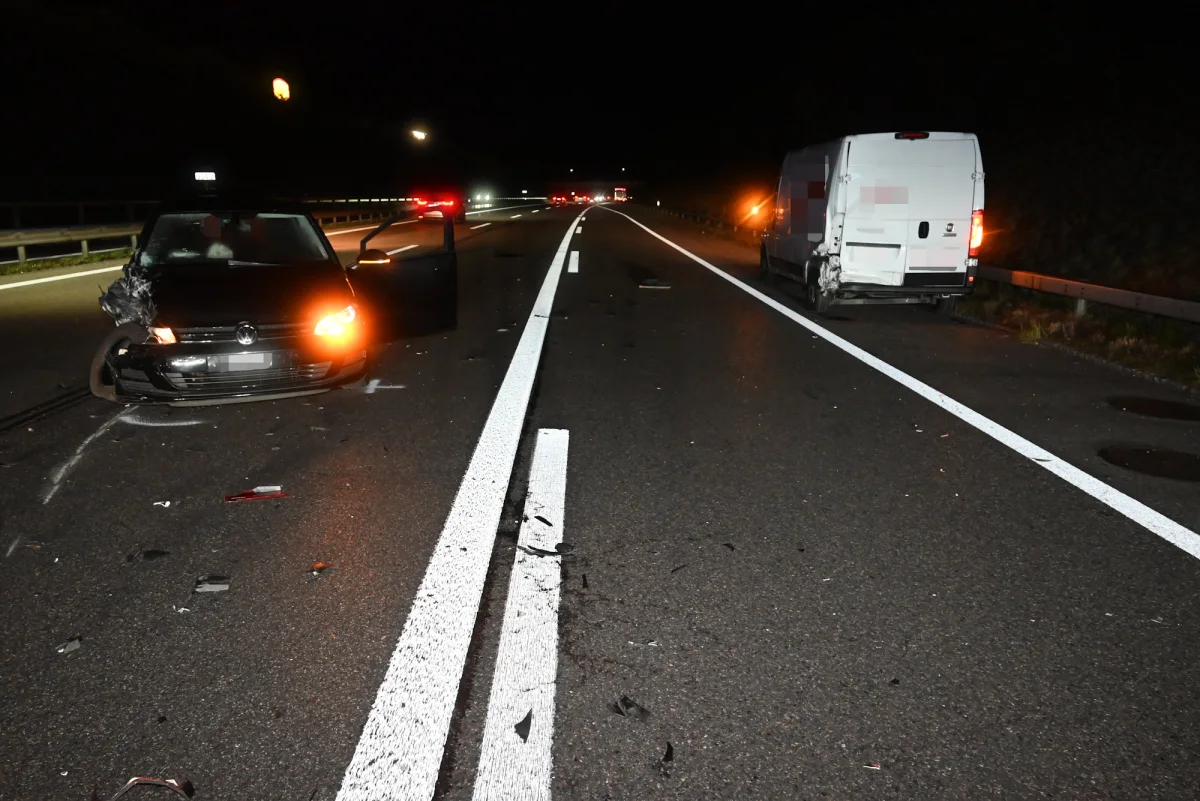 Ein schwarzer PKW mit Frontschaden steht auf einer Autobahn, daneben ein weißer Transporter mit Heckschaden. Es ist Nacht.