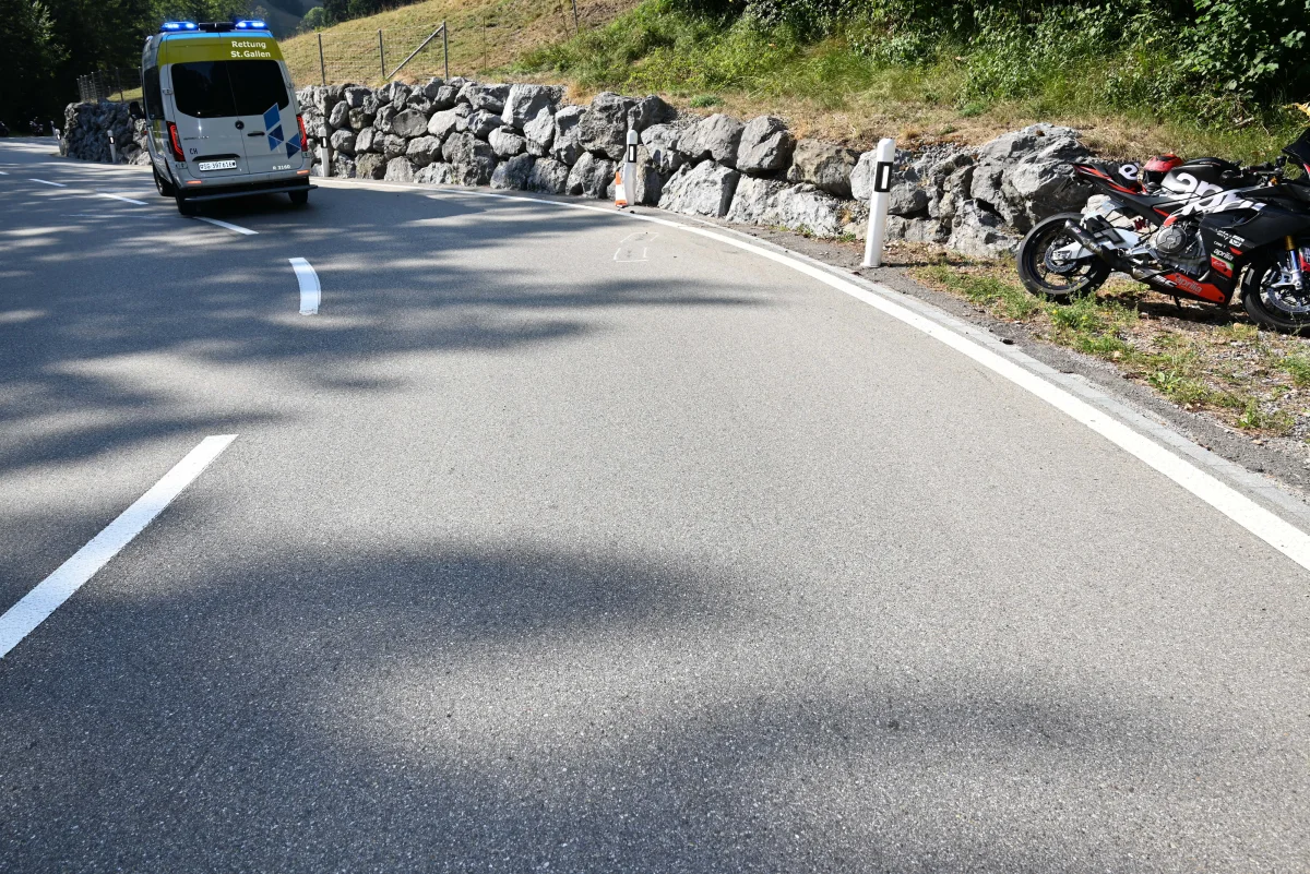 Auf einer kurvigen Landstraße ist ein gelber Rettungswagen von St. Gallen sichtbar. Am rechten Straßenrand steht ein schwarzes Motorrad neben einer Steinmauer.