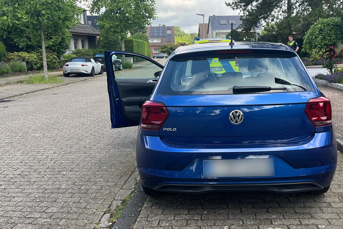 Ein blauer VW Polo steht mit geöffneter Fahrertür auf einer Wohnstraße. Im Hintergrund ist ein Polizeifahrzeug zu sehen.