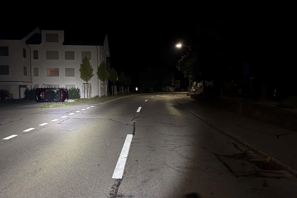 Ein Auto liegt umgestürzt auf einer nächtlichen Wohnstraße in der Nähe eines Gebäudes. Straßenbeleuchtung ist aktiv.