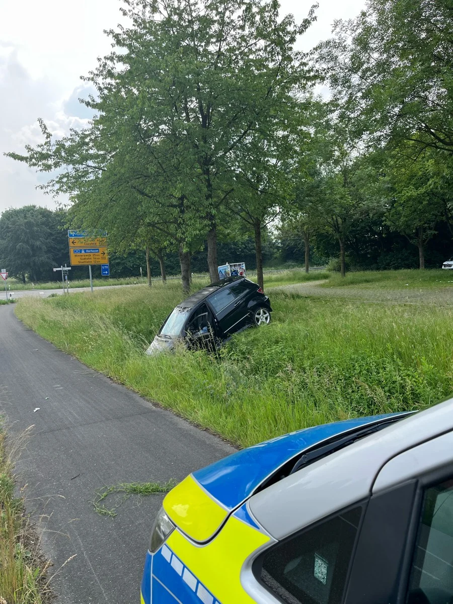 Ein Auto ist seitlich in einen Graben gefahren und steht schräg. Im Vordergrund ist ein Teil eines Polizeifahrzeugs sichtbar.