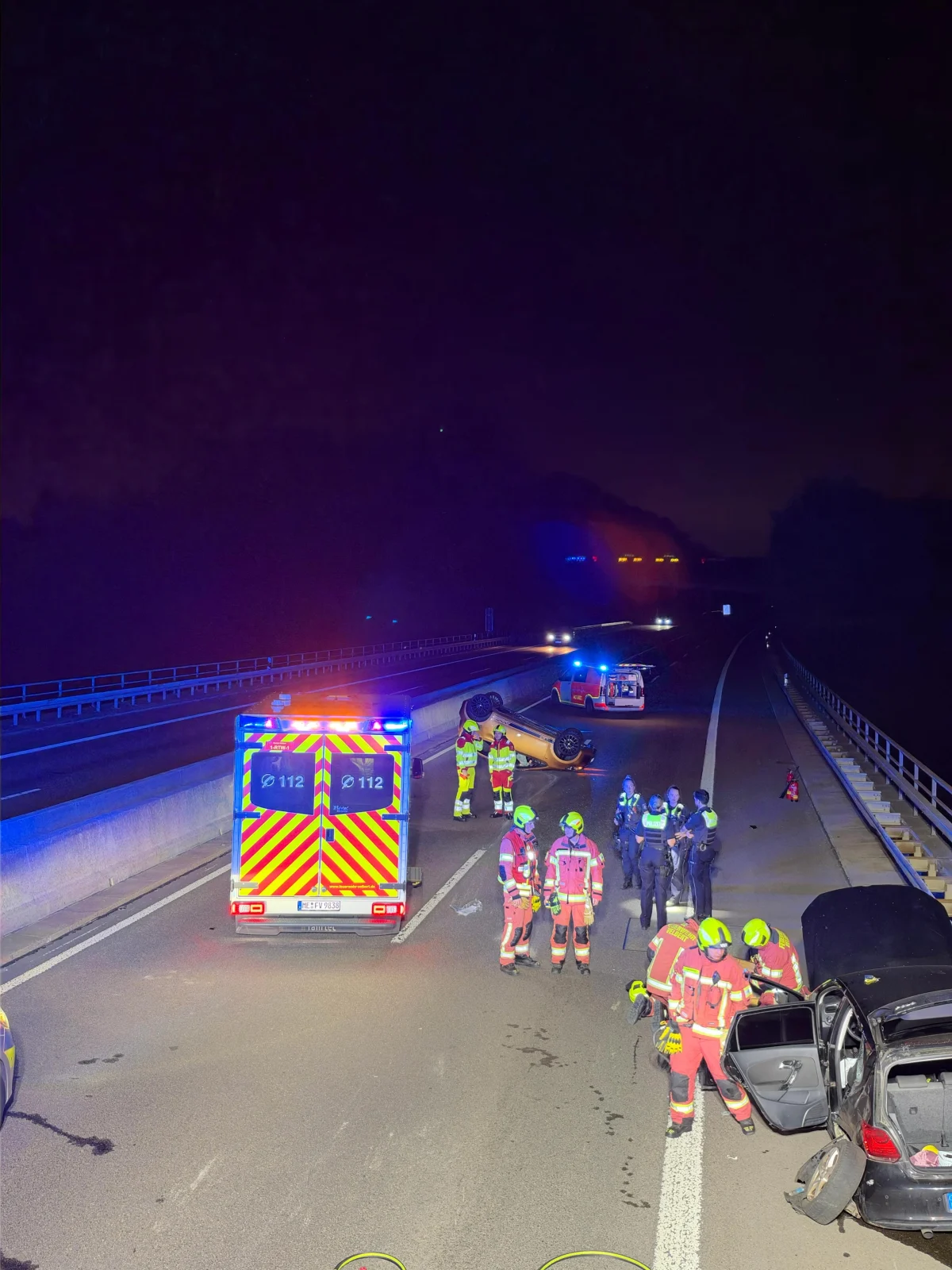 Ein umgestürztes Auto liegt auf einer Autobahn bei Nacht. Rettungskräfte in farbigen Westen sind vor Ort, um die Lage zu sichern.