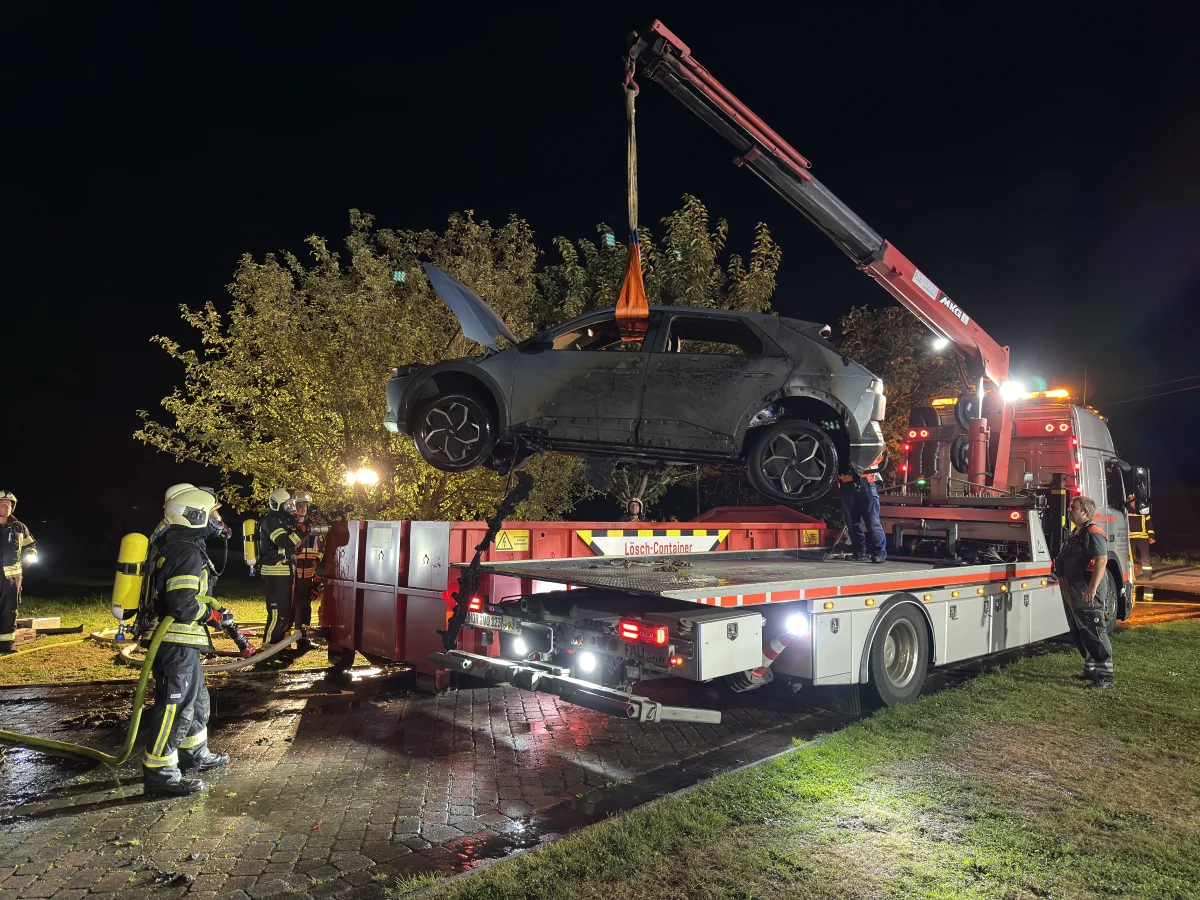 Mehrere Feuerwehrleute stehen um ein ausgebranntes Auto, das von einem Kran auf einen Abschleppwagen gehoben wird. Es ist Nacht.