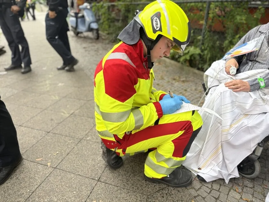 Ein Rettungssanitäter in gelb-roter Uniform kniet auf dem Gehweg und betreut eine Person im Rollstuhl mit einem Infusionszugang am Arm.
