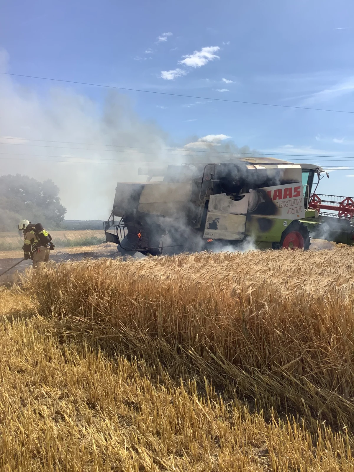 Ein Mähdrescher steht auf einem Feld in Flammen, während ein Feuerwehrmann versucht, das Feuer unter Kontrolle zu bringen. Rauch steigt in die Luft, es ist sonnig.
