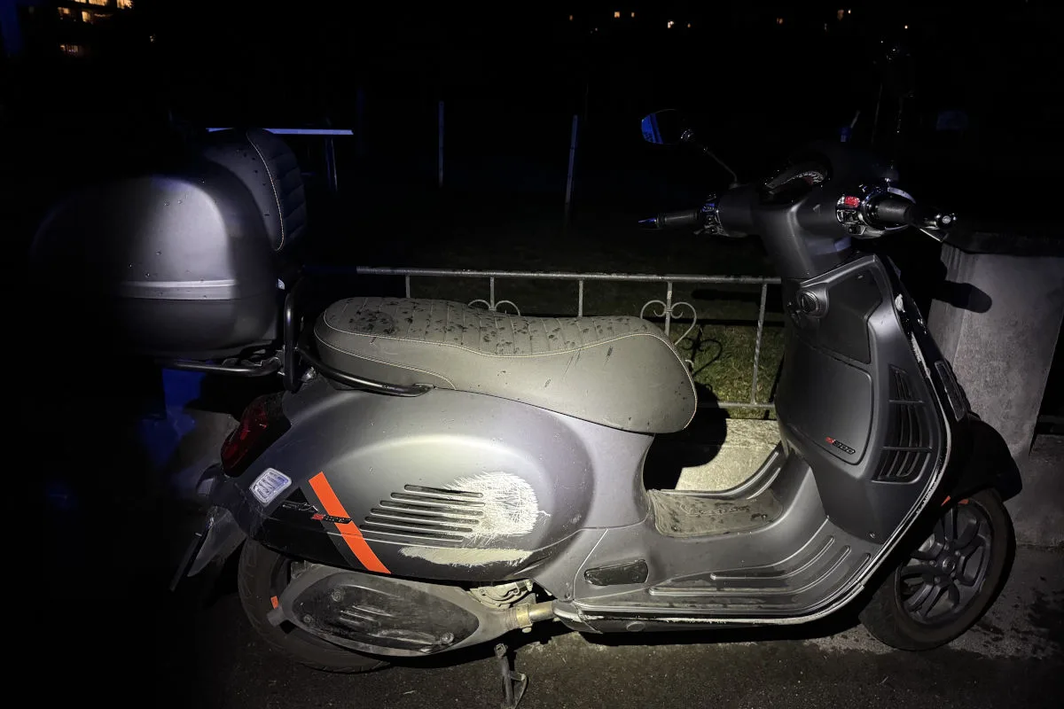 Ein grauer Roller mit sichtbaren Kratzern und Schmutz steht draußen im Dunkeln. Der Bereich ist schwach beleuchtet.