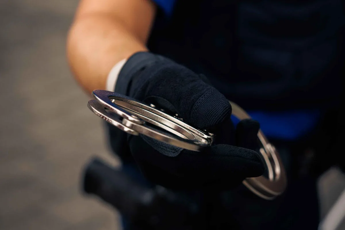 Ein Polizist mit Handschuhen hält ein Paar Handschellen in der Hand, bereit für den Einsatz. Der Hintergrund ist unscharf.