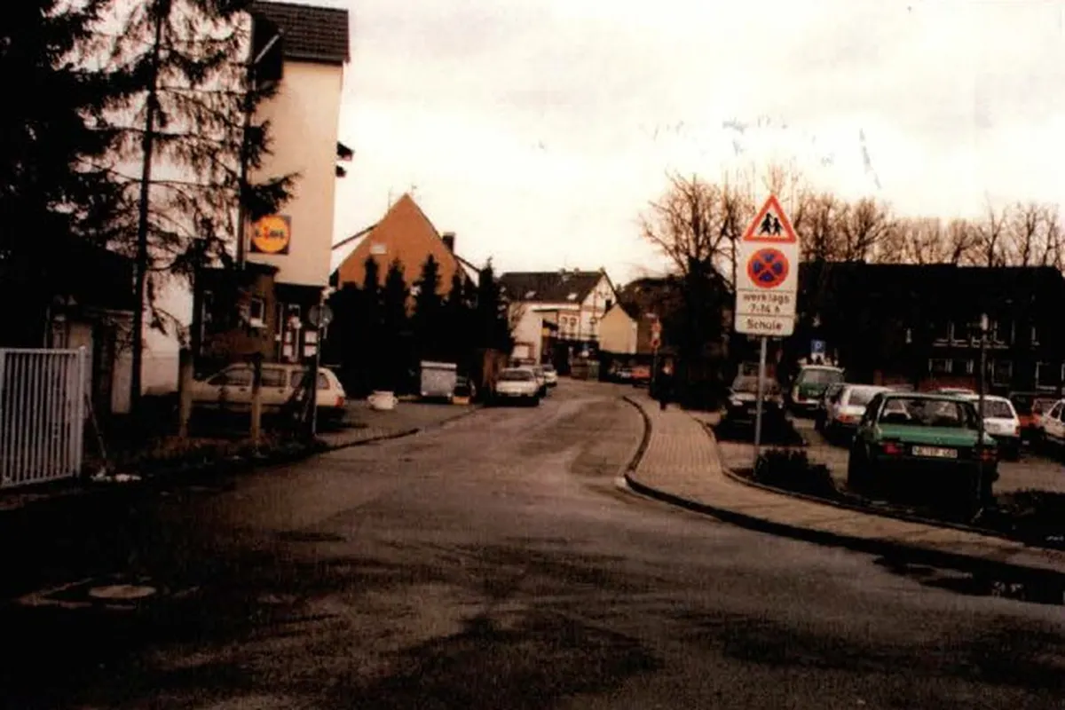 Das Bild zeigt eine Wohnstraße mit mehreren parkenden Autos und Verkehrsschildern im Vordergrund. Die Straße ist leicht gebogen und Bäume sind im Hintergrund sichtbar.