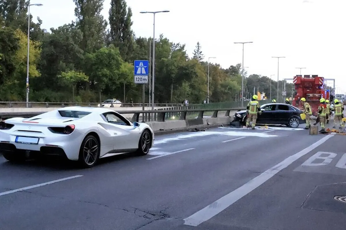 Ein weißer Sportwagen steht auf der Autobahn. Im Hintergrund sichern Feuerwehrleute ein beschädigtes schwarzes Auto neben der Leitplanke.