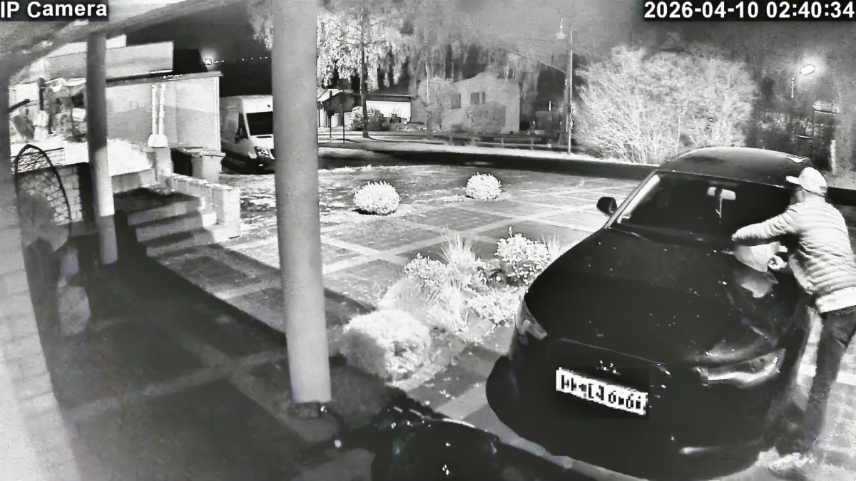 Eine Person lehnt sich über die Motorhaube eines Autos in einer nächtlichen Umgebung. Die Szene wird von einer Überwachungskamera aufgenommen.