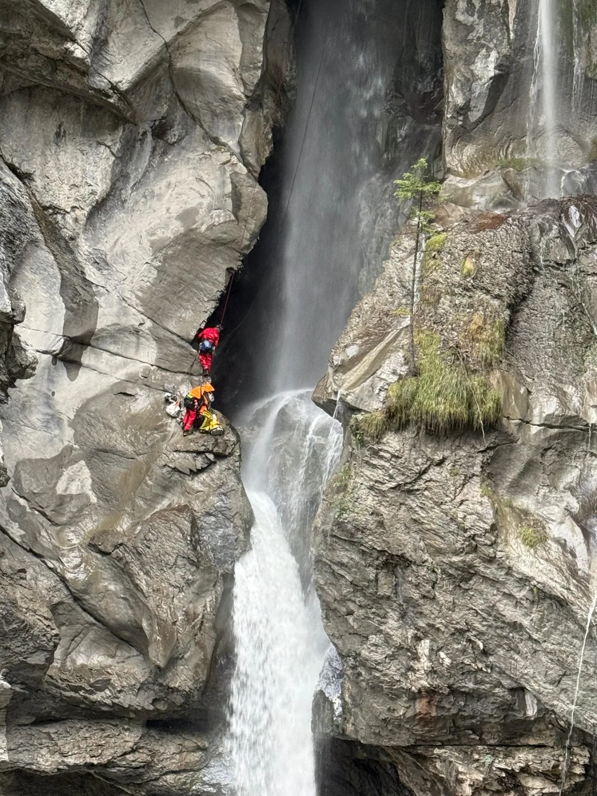 Zwei Rettungskräfte in leuchtenden Anzügen seilen sich an einer steilen Felswand neben einem Wasserfall ab.