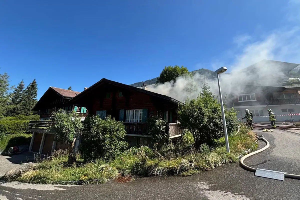 Ein Chalet mit Rauchentwicklung am Dach, zwei Feuerwehrleute sind im Einsatz und ein Schlauch liegt am Boden.