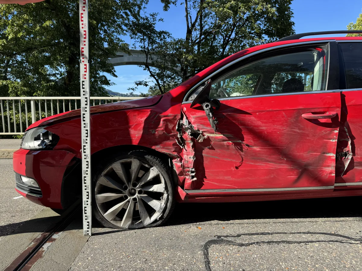 Ein rotes Auto mit erheblichen Schäden an der rechten Seite. Ein Maßstab ist vertikal daneben platziert. Der Hintergrund zeigt Bäume und eine Brücke.