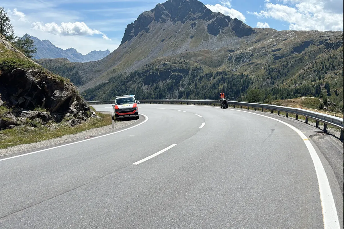 Ein Polizeiauto ist auf einer kurvigen Bergstraße in einer alpinen Umgebung geparkt. Ein Polizist in einer Warnweste steht daneben.