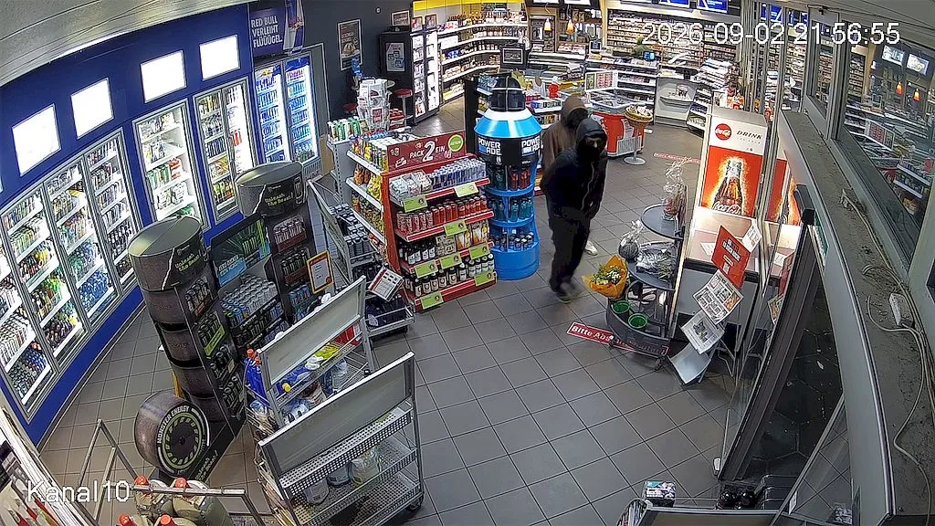 Innenaufnahme eines Ladenbereichs mit Regalen, gekühlten Getränken und zwei Personen, die durch den Gang gehen.