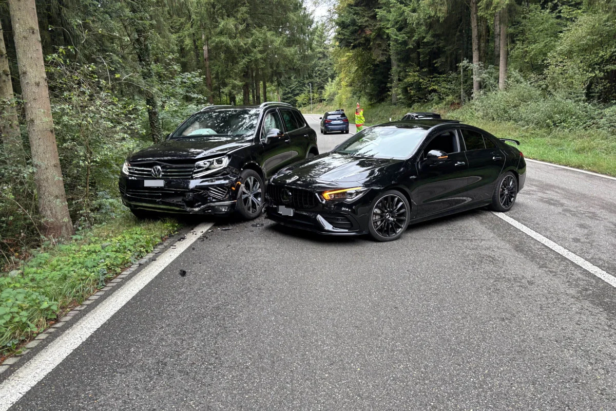 Auf einer Landstraße vor einem Wald sind zwei schwarze Autos frontal kollidiert. Ein Mitarbeiter in Warnweste ist im Hintergrund sichtbar.