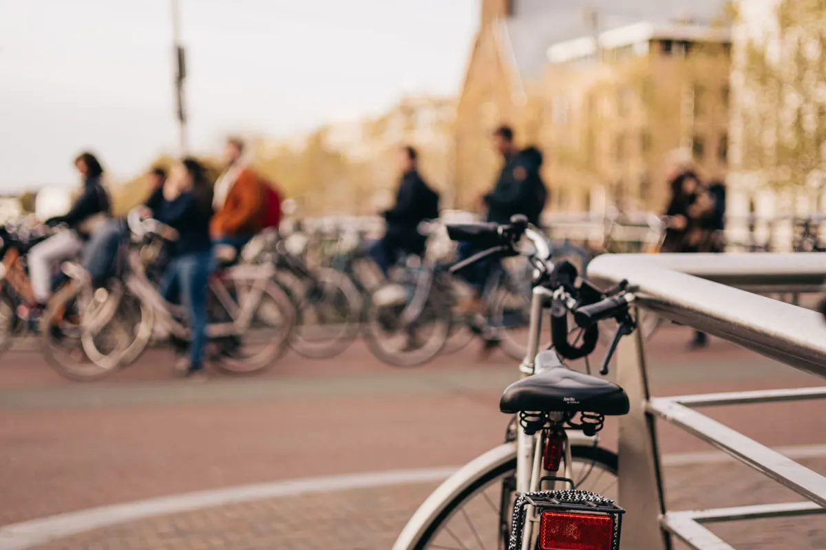 Im Vordergrund lehnt ein Fahrrad an einem Geländer, während im Hintergrund unscharf mehrere Personen auf Fahrrädern vorbeifahren. Die Szene scheint in einer städtischen Umgebung bei Tageslicht aufgenommen worden zu sein.