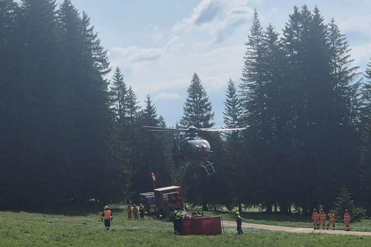 Ein Hubschrauber holt Wasser aus einem Behälter auf einer Wiese nahe einem Wald. Personen in orangenen Westen sind im Hintergrund sichtbar.