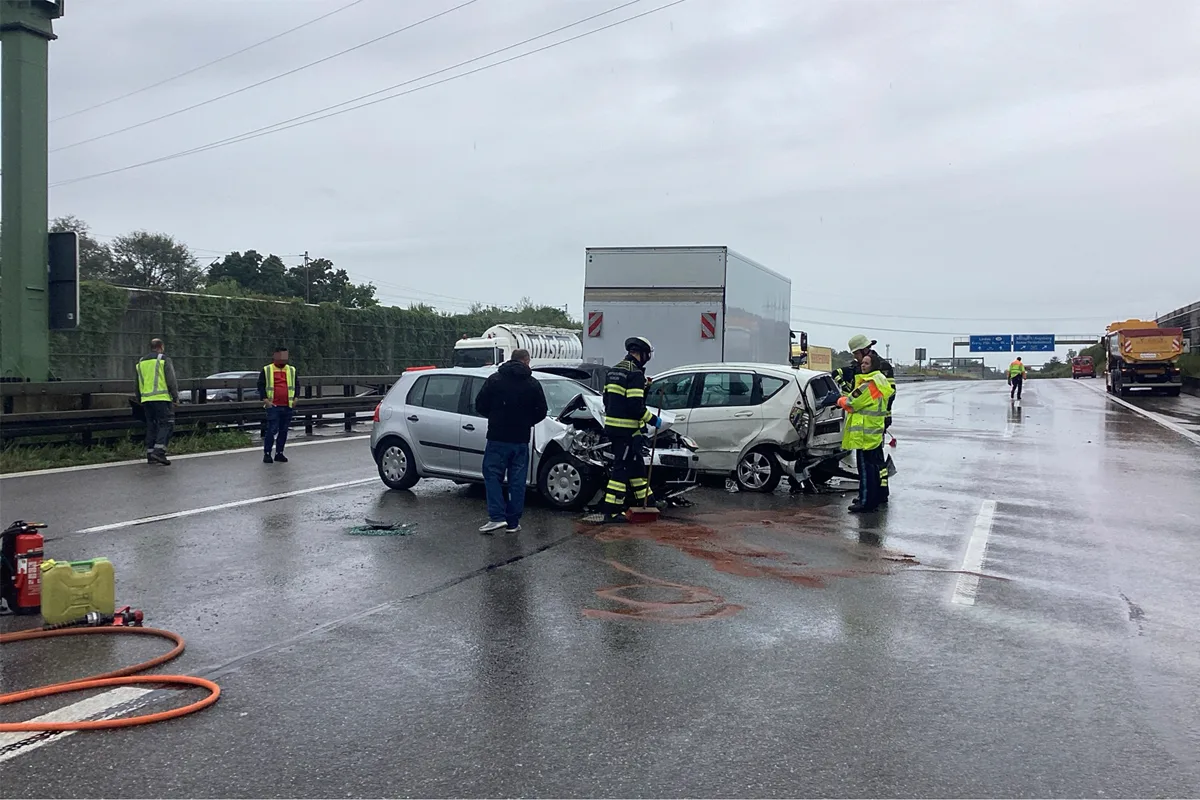 Auf einer nassen Autobahn sind zwei Fahrzeuge zusammengestoßen. Feuerwehrleute arbeiten an der Unfallstelle, während ein Lastwagen im Hintergrund steht.