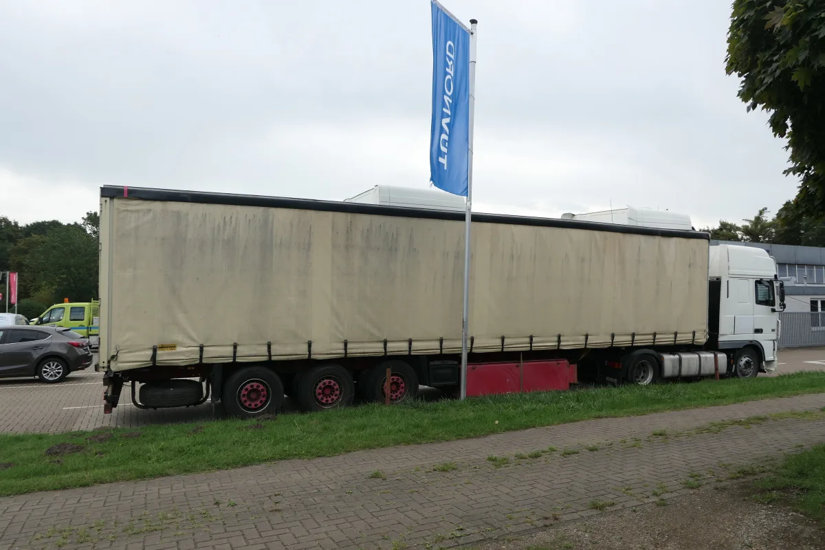 Ein weißer Lastwagen mit roter Felge steht geparkt auf einem gepflasterten Parkplatz. Im Hintergrund sind Bäume und ein Firmengebäude zu erkennen.