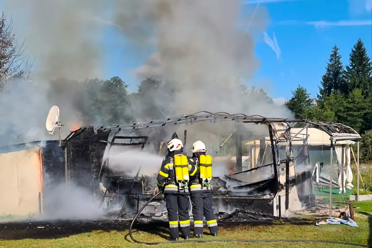 Zwei Feuerwehrleute in Schutzanzügen löschen einen brennenden Wohnwagen. Starker Rauch steigt auf.
