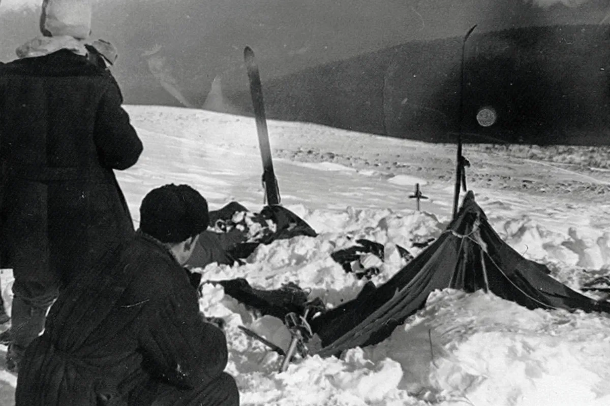 Das Bild zeigt ein verschneites Zelt mit Skiern daneben in einer winterlichen Landschaft. Mehrere Personen stehen oder hocken im Schnee um das Zelt herum.