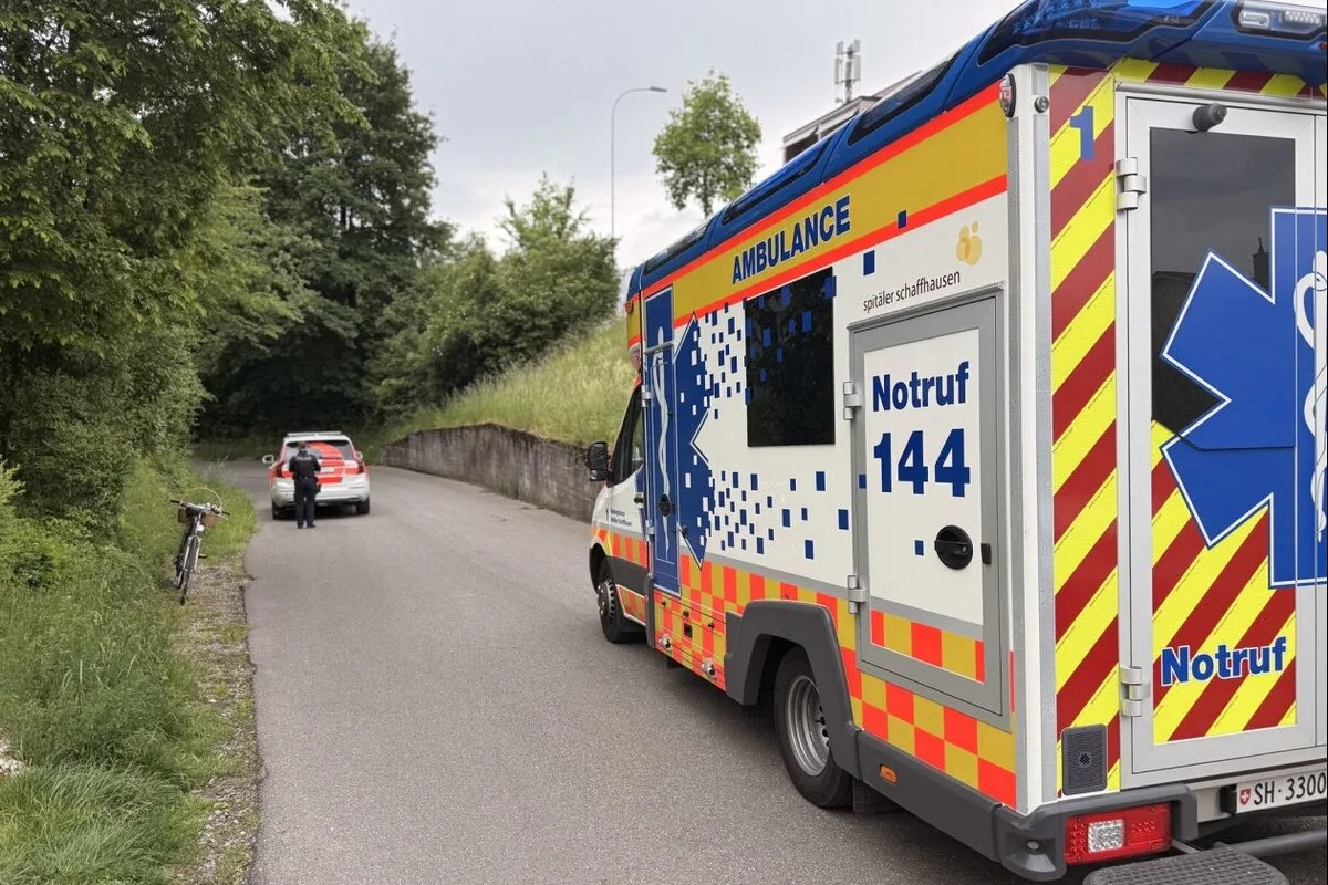 Ein Rettungswagen mit der Aufschrift 'Notruf 144' und ein Polizeifahrzeug stehen auf einer ruhigen, bewaldeten Straße. Ein Fahrrad lehnt am Straßenrand.