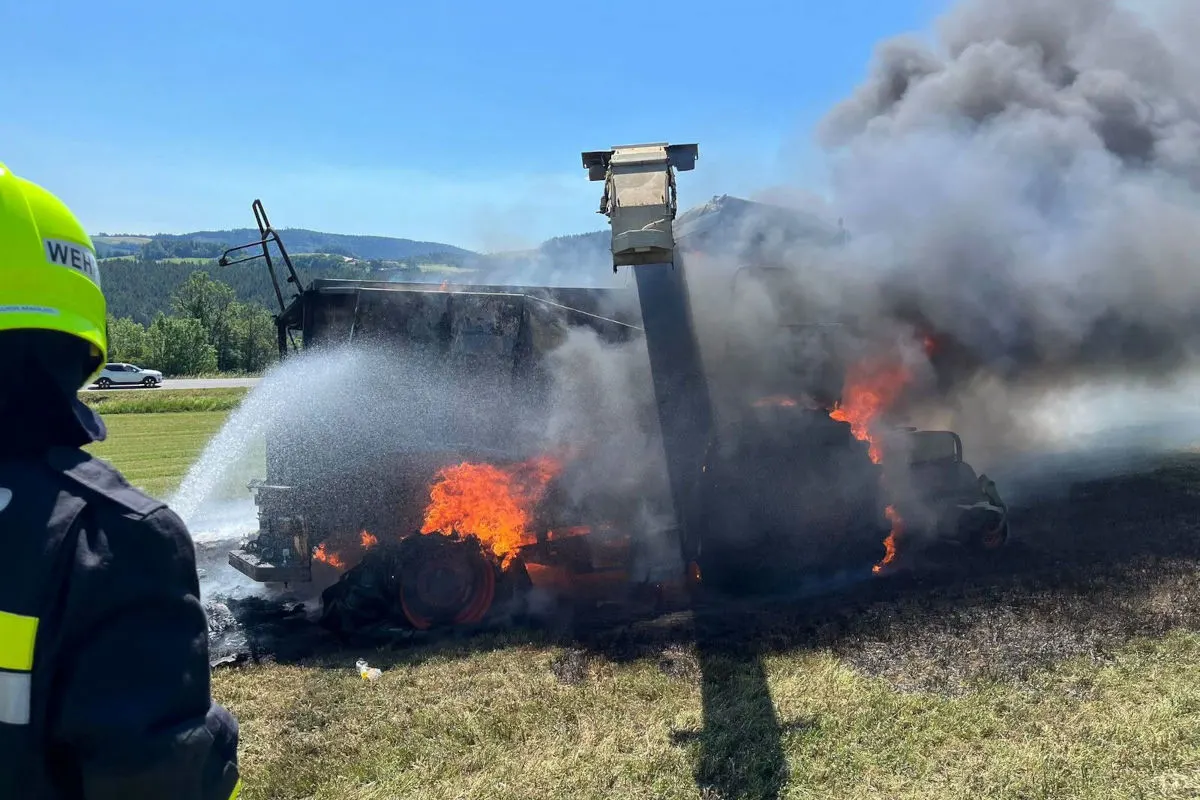 Ein brennender Lastwagen wird auf einem Feld von der Feuerwehr mit einem Wasserschlauch gelöscht. Rauch steigt auf.