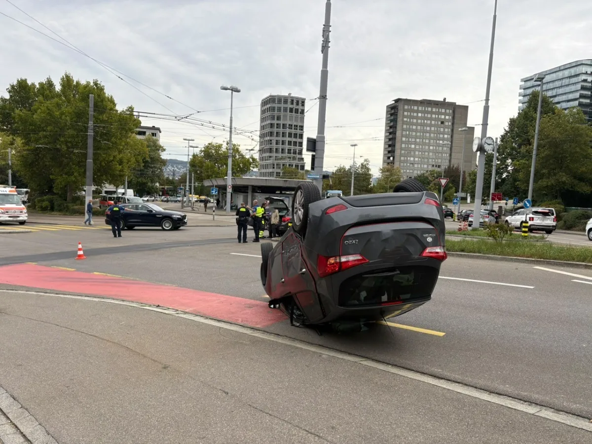 Auf dem Bild liegt ein Auto auf dem Dach mitten auf einer großen innerstädtischen Kreuzung. Im Hintergrund sind mehrere Gebäude sowie Einsatzkräfte der Polizei sichtbar.