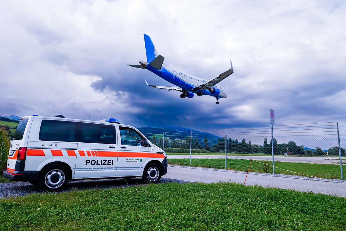 Ein Polizeifahrzeug der Kantonspolizei St. Gallen steht nahe einer Landebahn, während ein Flugzeug darüber fliegt. Der Himmel ist bewölkt, und im Hintergrund sind Felder und Hügel sichtbar.